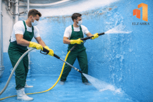 شركة عزل وتنظيف الخزانات بحي الغويلا – نجران عاملان يقومان بتنظيف الخزان باستخدام معدات متخصصة وخراطيم المياه مع ظهور لوجو الزهراء أعلى يمين الصورة